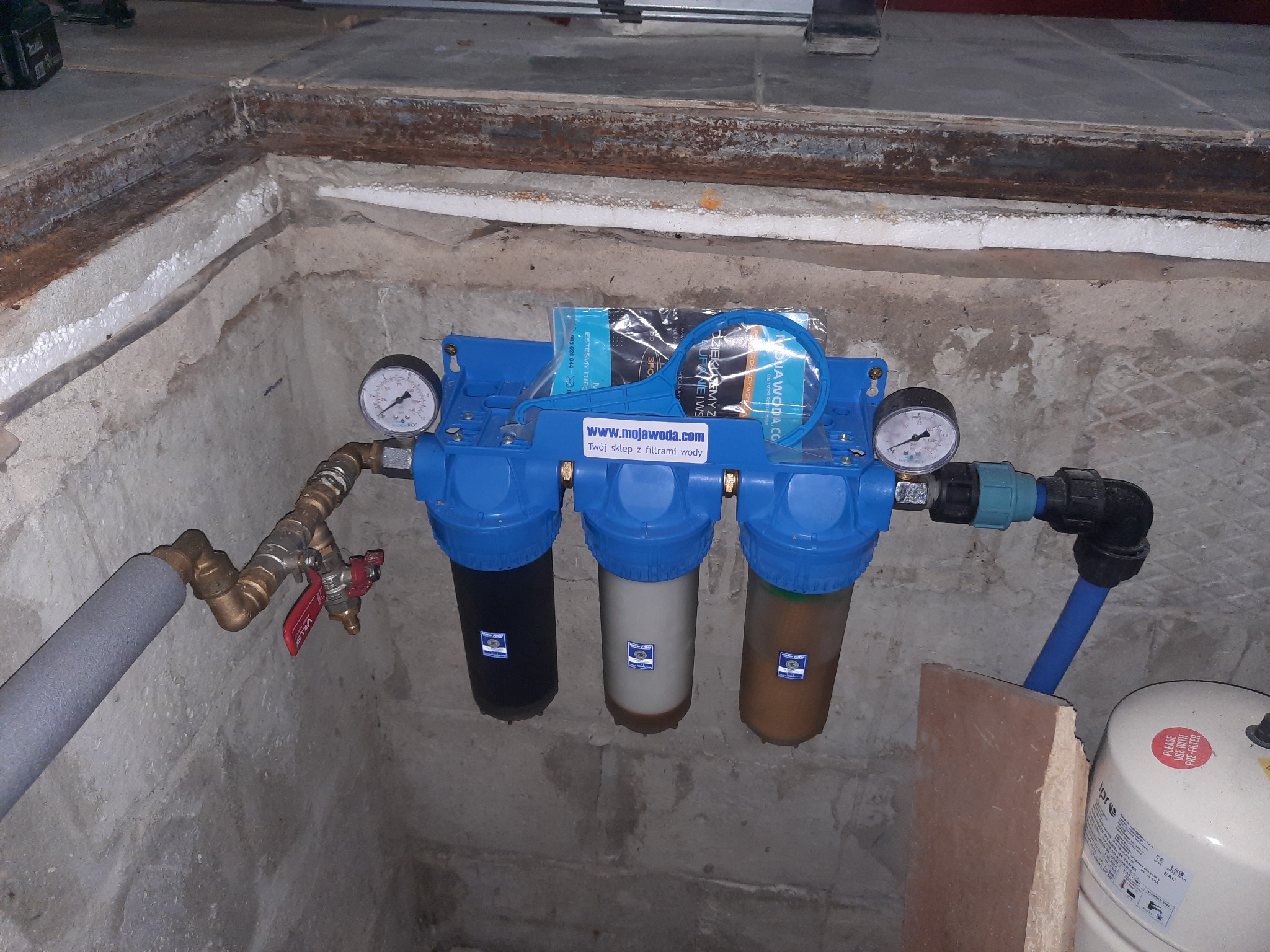 Trzyczęściowy filtr wody z manometrami, zainstalowany w surowym pomieszczeniu z betonowymi ścianami i rurami. Widoczne filtry o różnym stopniu zabrudzenia i akcesoria montażowe. Na górze filtra...