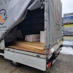 TRANS - PIOTR RYBA TRANSPORT I PRZEPROWADZKI - Żółty samochód dostawczy firmy Trans-Piotr z częściowo odsłoniętą plandeką, załadowany płytami i materiałami izolacyjnymi, na tle składu budowlanego z paletami materiałów w zimowej scenerii.