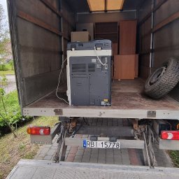TRANS - PIOTR RYBA TRANSPORT I PRZEPROWADZKI - Wnętrze samochodu dostawczego z załadowanym biurowym sprzętem, w tym dużą drukarką i szafką, podczas przeprowadzki, widoczna opona i fragment tablicy rejestracyjnej.