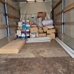 TRANS - PIOTR RYBA TRANSPORT I PRZEPROWADZKI - Wnętrze paki samochodu dostawczego wypełnione kartonami, meblami i innymi przedmiotami gotowymi do transportu. Widoczne płyty meblowe i profile aluminiowe.