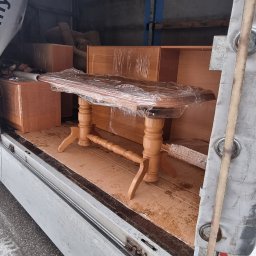 TRANS - PIOTR RYBA TRANSPORT I PRZEPROWADZKI - Drewniany stół owinięty folią stretch wewnątrz brudnej przestrzeni ładunkowej samochodu dostawczego, obok kartonów i regału.