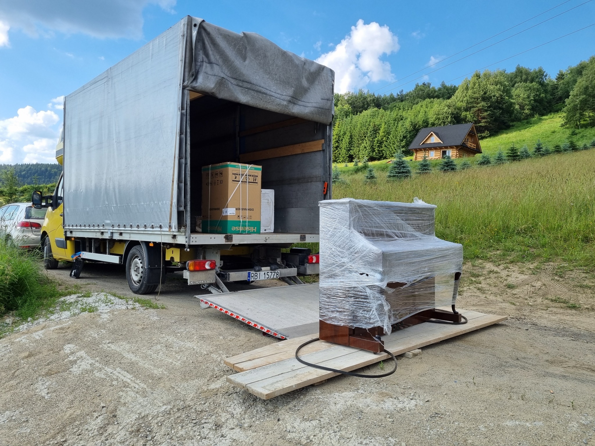 Pianino owinięte folią stretch na drewnianych deskach, przygotowane do załadunku na platformę samochodu dostawczego w plenerze z widokiem na drewniany dom na wzgórzu.