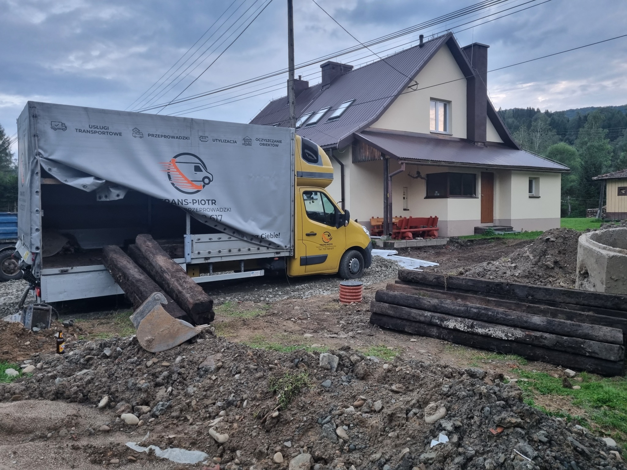 Żółta furgonetka firmy transportowej z częściowo otwartą plandeką, z której wysuwają się ciemne, drewniane bale, zaparkowana na terenie budowy przed domem jednorodzinnym; obok sterta ziemi z łopatą...