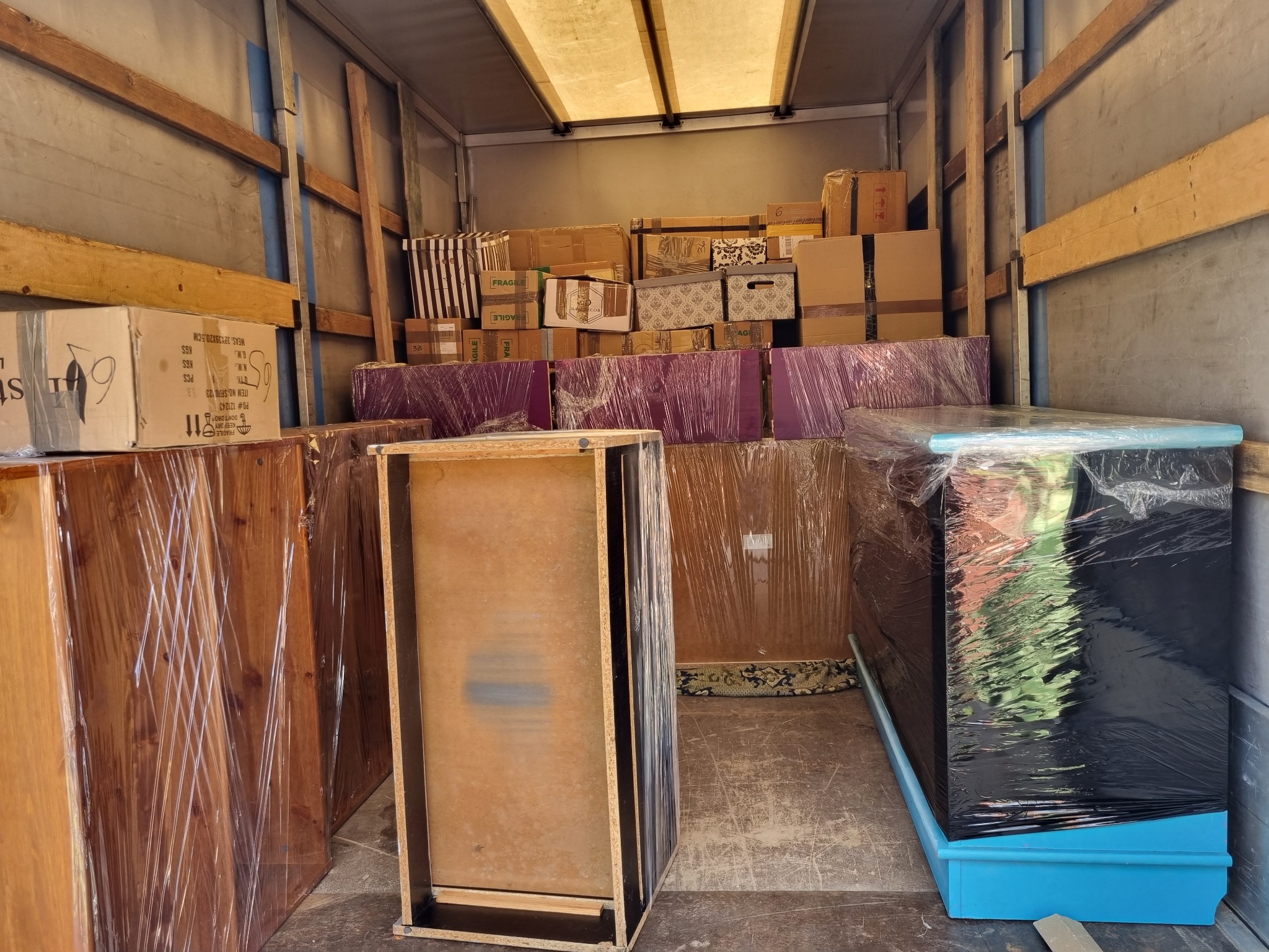 Wnętrze furgonetki wypełnionej kartonami i meblami zabezpieczonymi folią stretch, gotowymi do transportu. Widoczny fragment drewnianej szafki bez drzwiczek oraz zapakowane komody w różnych kolorach.