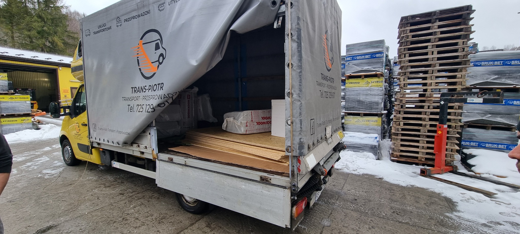 Żółty samochód dostawczy firmy Trans-Piotr z częściowo odsłoniętą plandeką, załadowany płytami i materiałami izolacyjnymi, na tle składu budowlanego z paletami materiałów w zimowej scenerii.