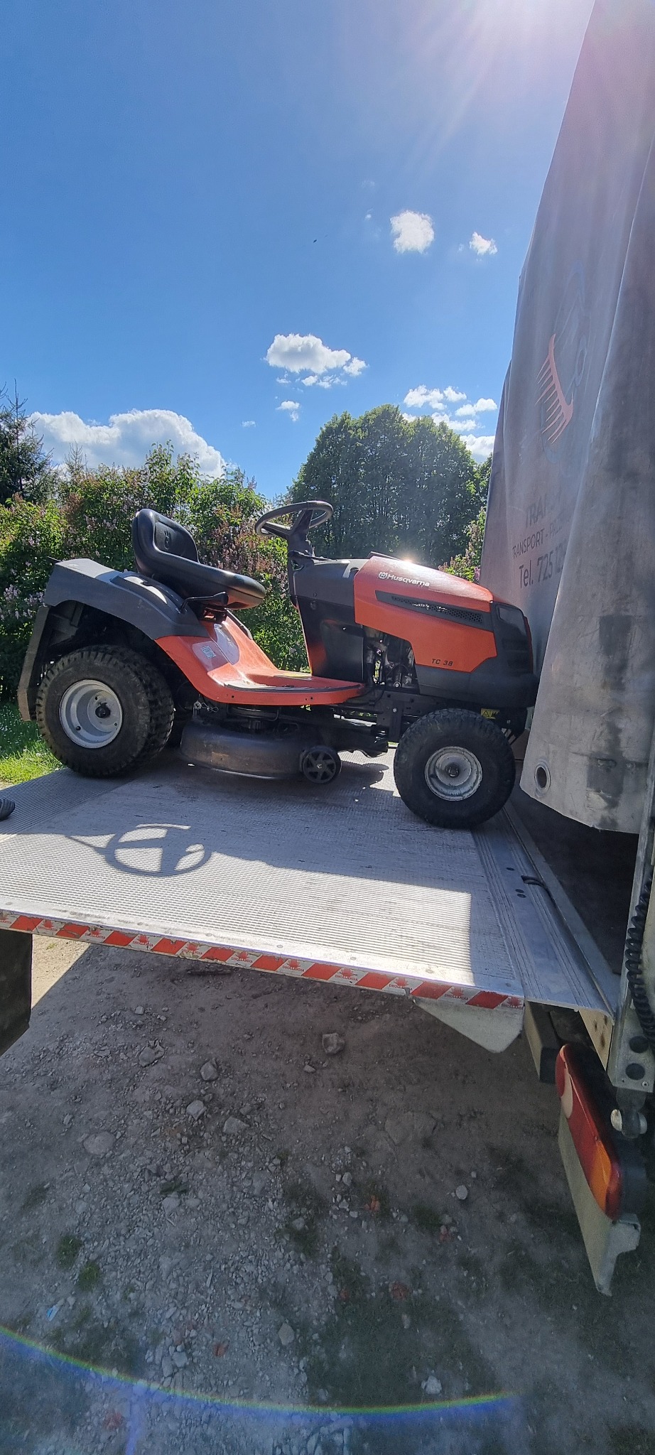 Pomarańczowy traktorek do koszenia trawy Husqvarna TC 38, załadowany na platformę samochodu transportowego z plandeką, w słoneczny dzień.