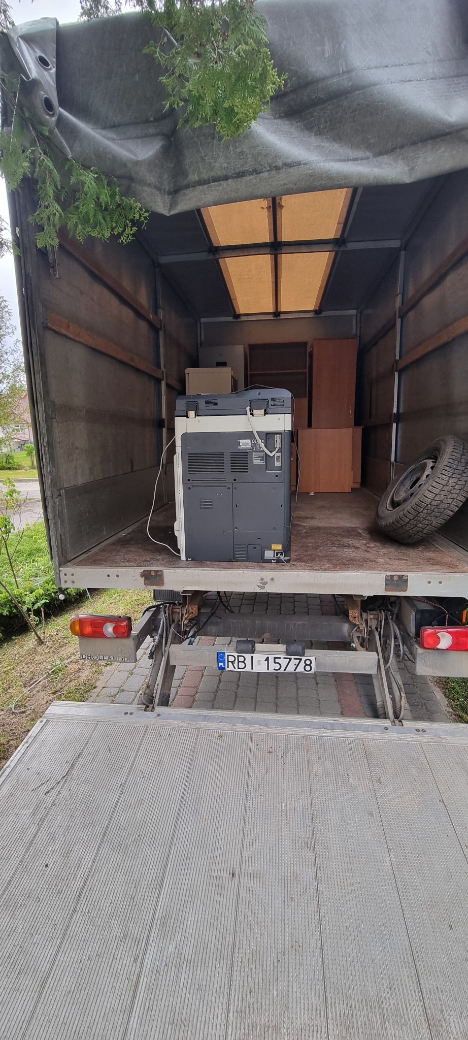 Wnętrze samochodu dostawczego z załadowanym biurowym sprzętem, w tym dużą drukarką i szafką, podczas przeprowadzki, widoczna opona i fragment tablicy rejestracyjnej.