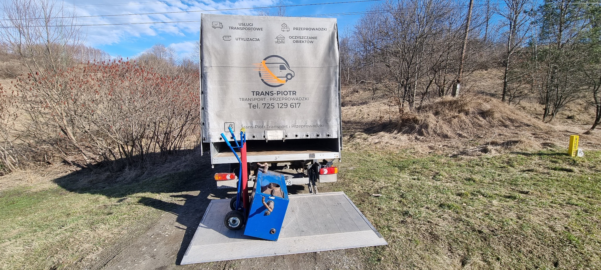 Tył samochodu dostawczego firmy TRANS-PIOTR z rampą załadowczą i wózkiem transportowym z niebieskim piecem na rampie, w tle widok na pagórkowaty teren z drzewami.