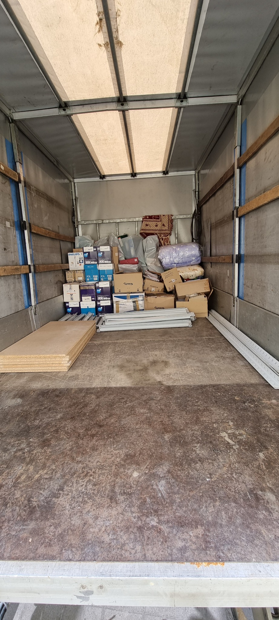 Wnętrze paki samochodu dostawczego wypełnione kartonami, meblami i innymi przedmiotami gotowymi do transportu. Widoczne płyty meblowe i profile aluminiowe.