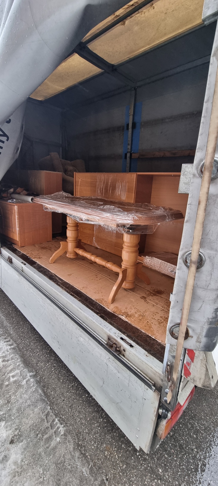 Drewniany stół owinięty folią stretch wewnątrz brudnej przestrzeni ładunkowej samochodu dostawczego, obok kartonów i regału.