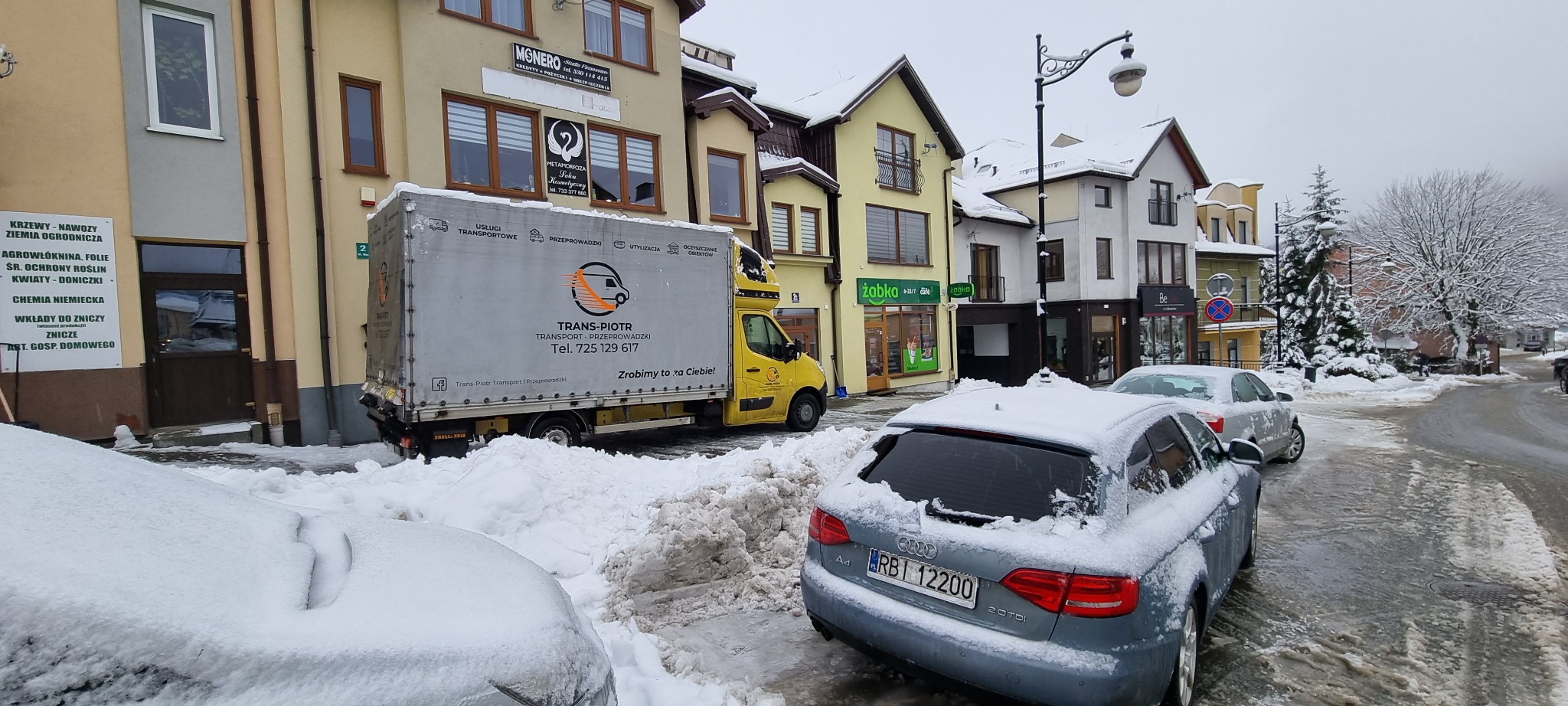 Żółta ciężarówka firmy Trans-Piotr z grafiką pomarańczowego samochodu w ruchu, zaparkowana na zasypanej śniegiem ulicy w zimowej scenerii miasteczka, w tle budynki z szyldami sklepów i usług.