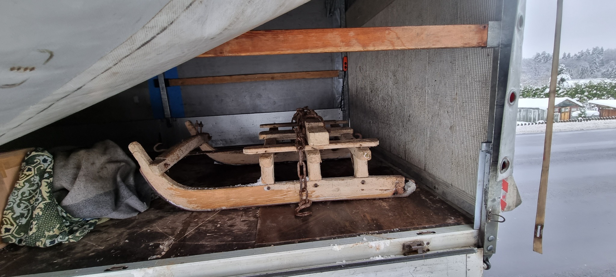 Drewniane sanie z metalowym łańcuchem, przewożone wewnątrz samochodu dostawczego, z widocznym śnieżnym krajobrazem za oknem.