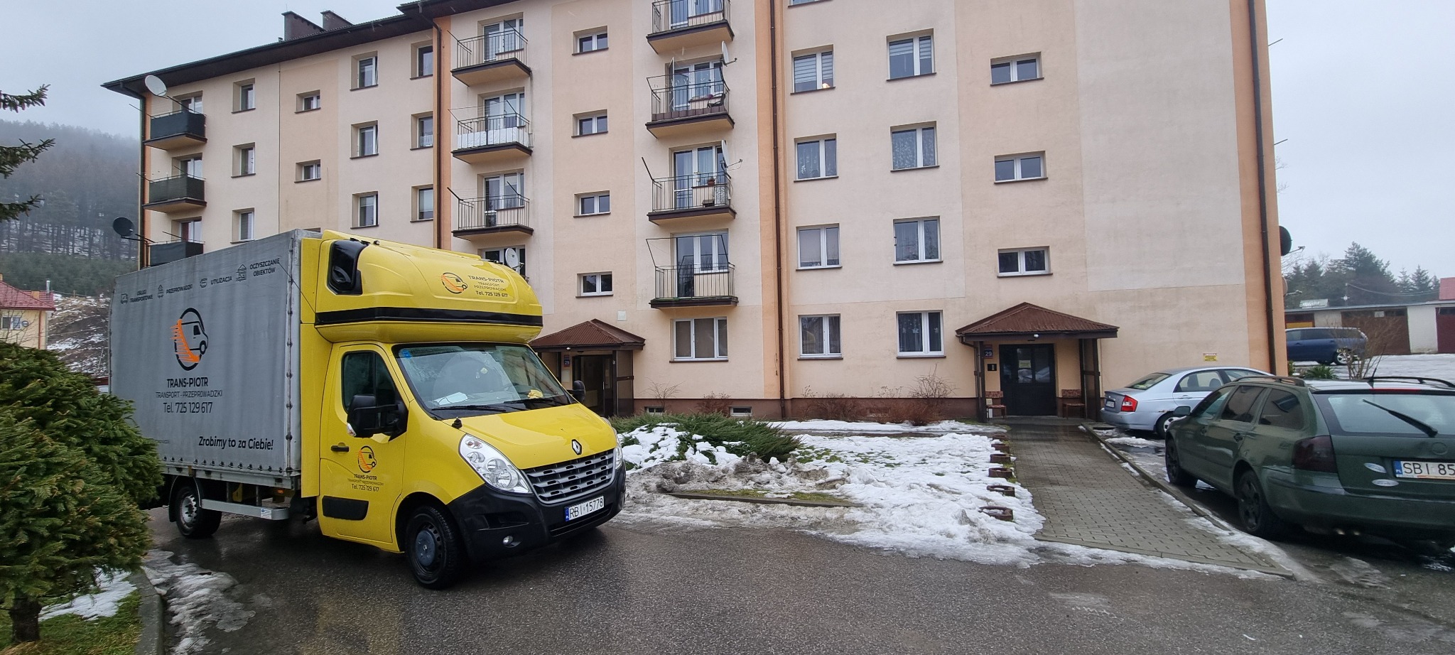Żółta furgonetka firmy transportowej TRANS-PIOTR zaparkowana przed budynkiem mieszkalnym z balkonami, widoczne resztki śniegu.