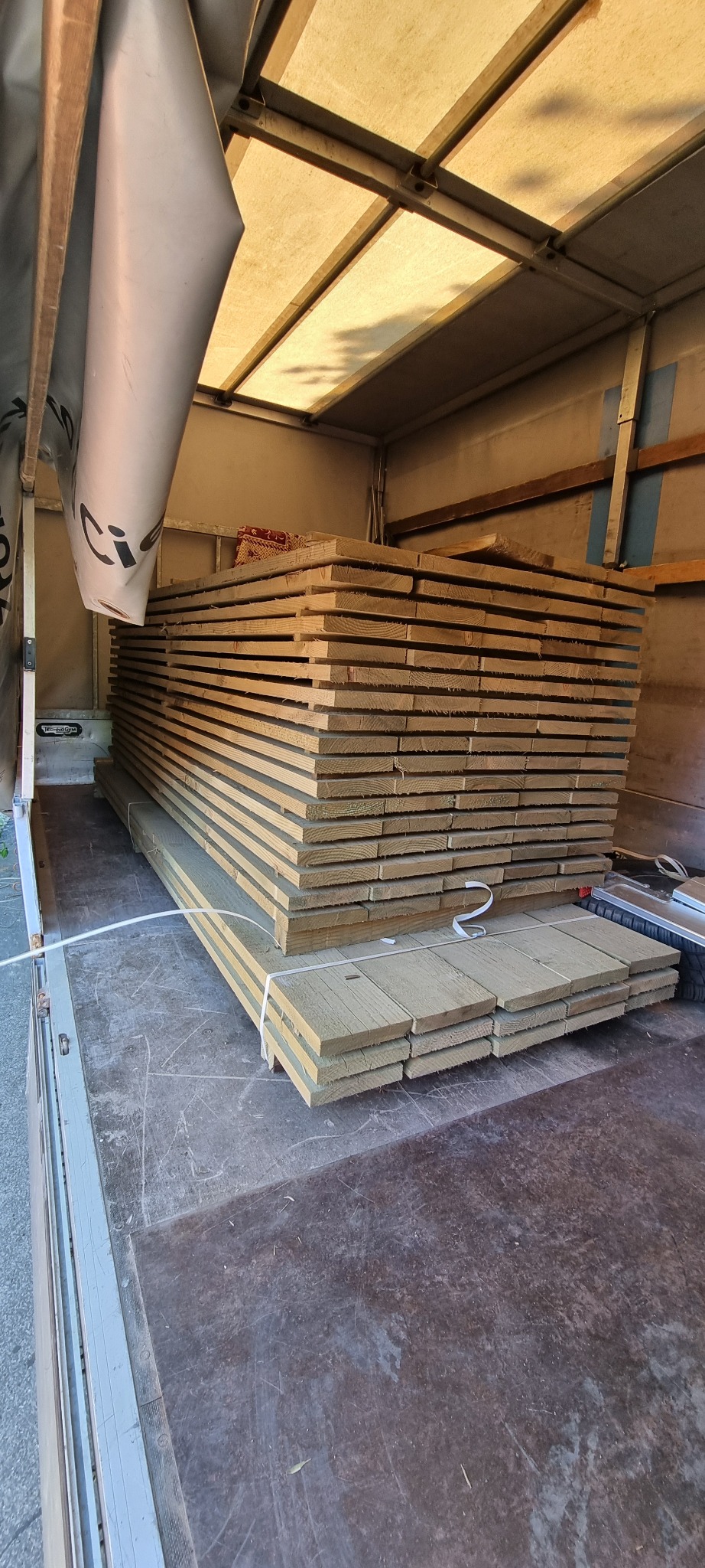 Stos desek budowlanych, zabezpieczonych białym sznurkiem, ułożony wewnątrz skrzyni samochodu dostawczego z rozpiętą plandeką.
