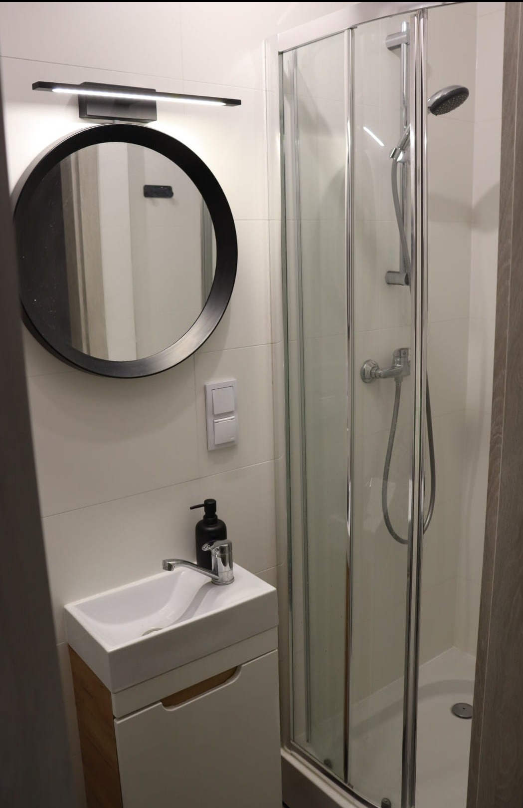 Nowoczesna, mała łazienka z kabiną prysznicową, okrągłym lustrem z czarną ramą, białą umywalką z szafką i czarnym dozownikiem mydła w płynie.