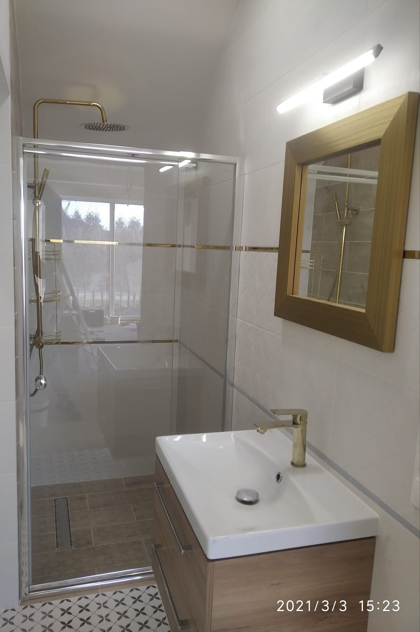 Nowoczesna łazienka z kabiną prysznicową typu walk-in z elementami w kolorze złotym, umywalką nablatową na drewnianej szafce i lustrem w złotej ramie, wykończona białymi płytkami z delikatnym wzorem.