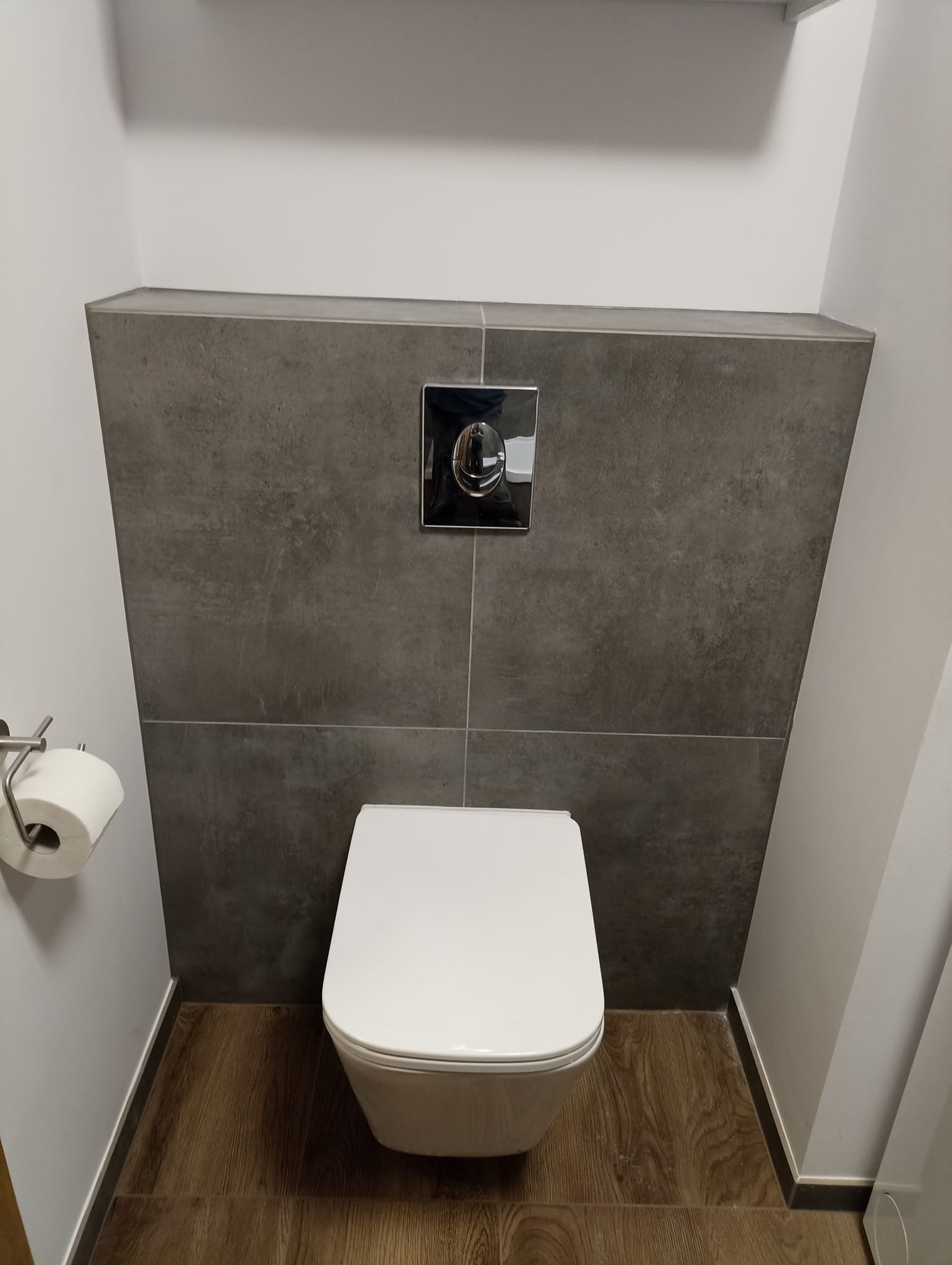 Podwieszana toaleta z chromowanym przyciskiem spłukującym na tle szarej ściany wyłożonej płytkami imitującymi beton, uzupełniona o drewnopodobną podłogę i uchwyt na papier toaletowy.