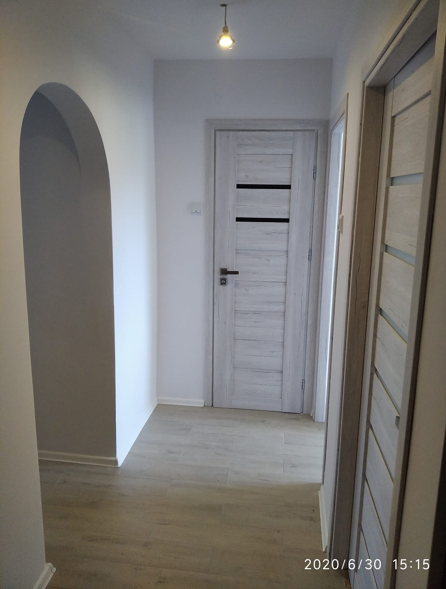 Korytarz po remoncie z dwiema parami drzwi w odcieniach bieli i szarości, podłoga imitująca drewno, oświetlenie punktowe sufitowe i łukowate przejście do innego pomieszczenia.