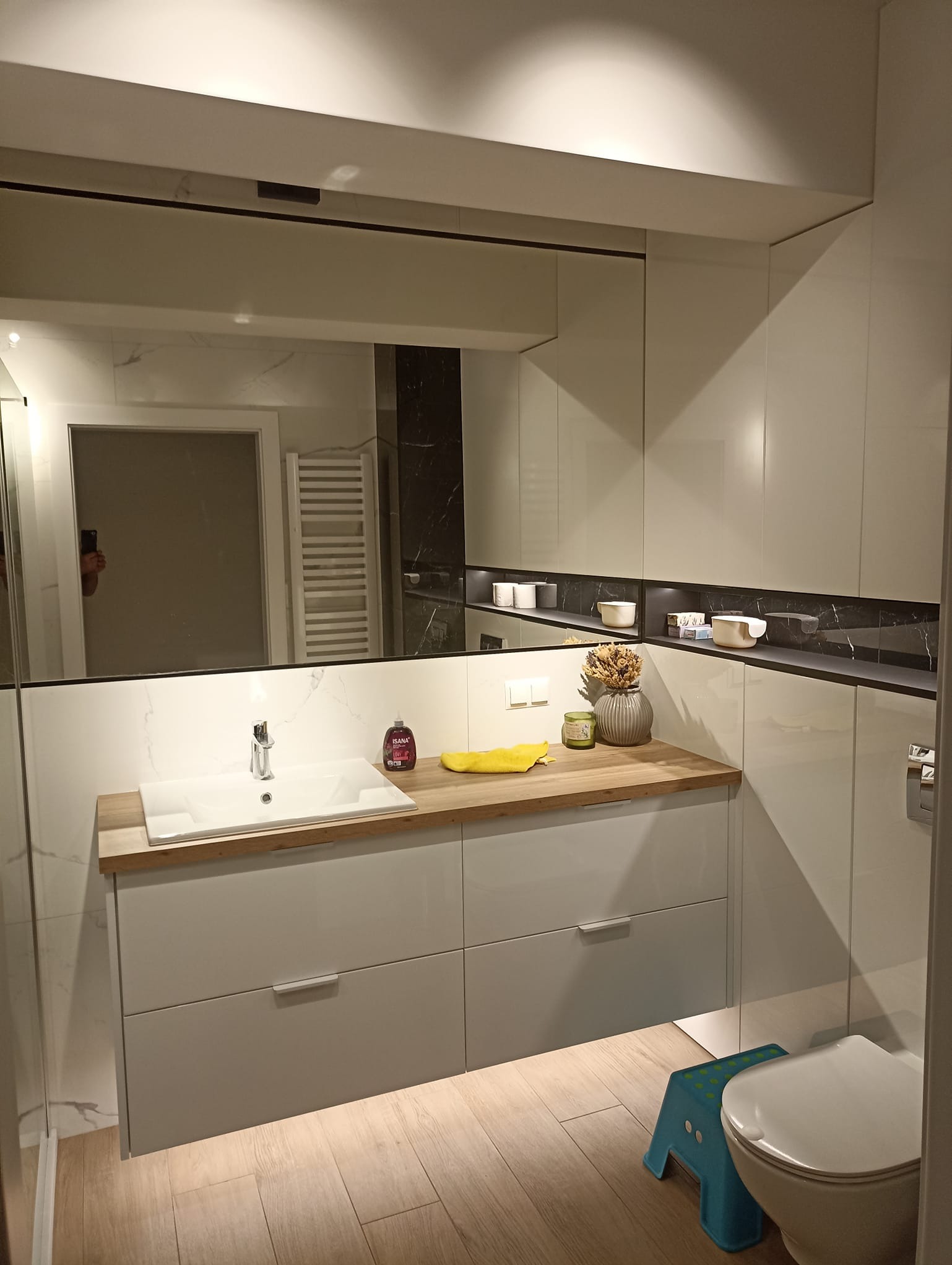 Nowoczesna łazienka z białymi szafkami pod umywalką z drewnianym blatem, duże lustro z odbiciem fragmentu łazienki, niebieski stołek dziecięcy obok toalety.
