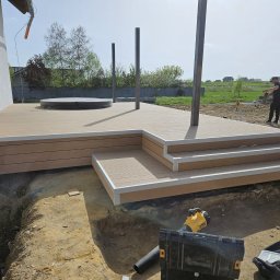 Zales - Montaż tarasu z widocznymi słupkami, schodami i przygotowanym podłożem. W tle operator minikoparki.