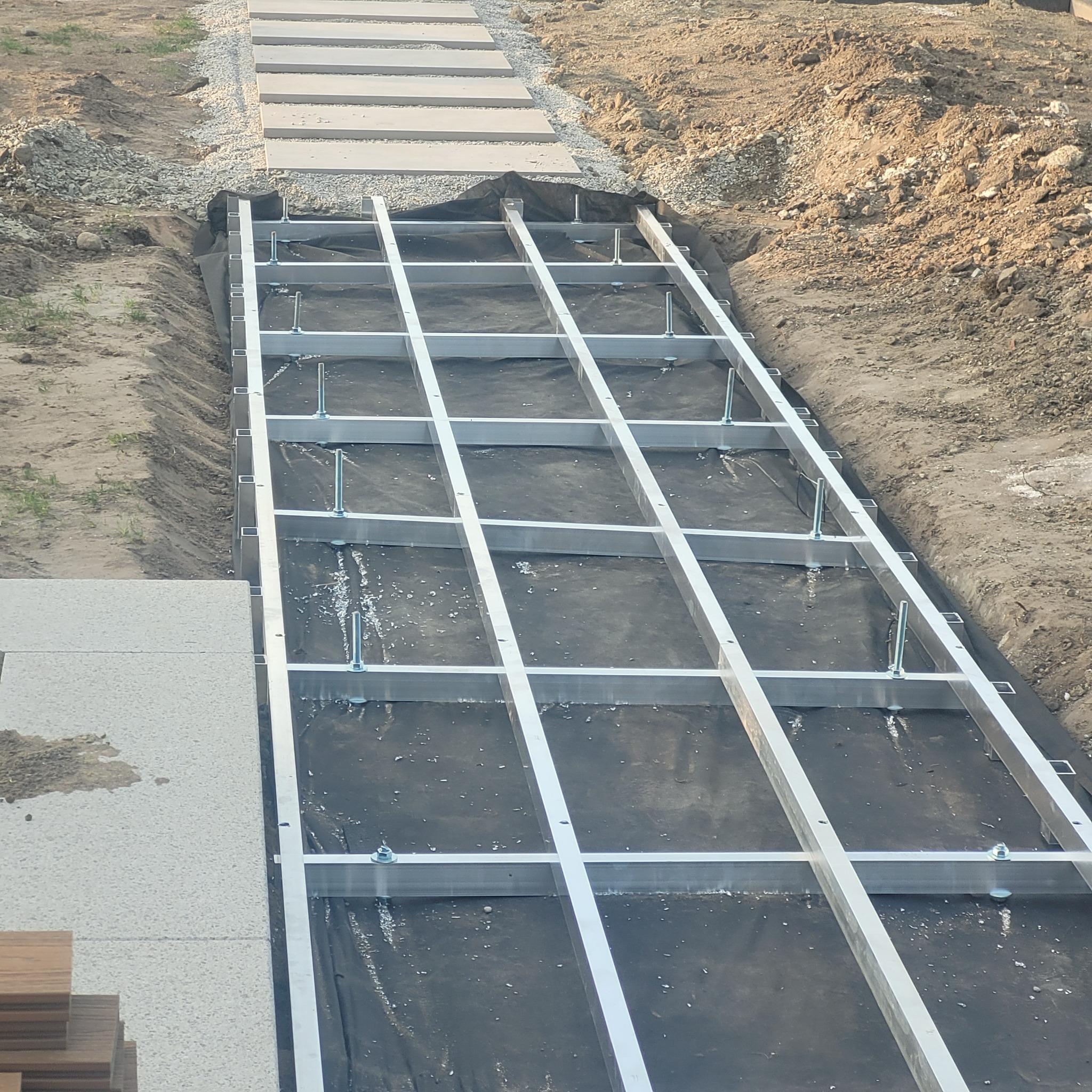 Konstrukcja aluminiowa pod przyszły taras, widoczne śruby mocujące i czarna membrana oddzielająca od gruntu. W tle ułożone płyty chodnikowe i przygotowane deski tarasowe.