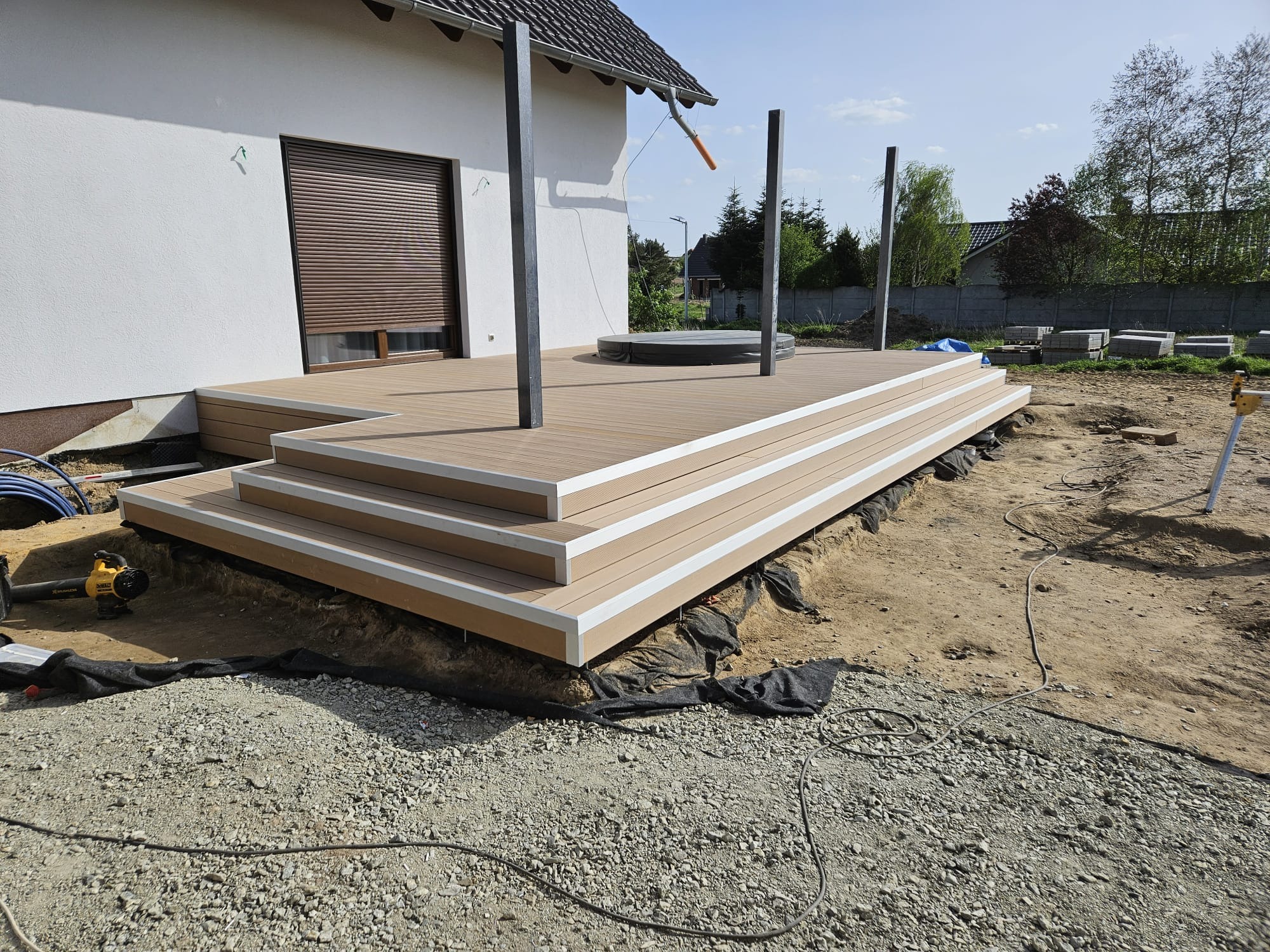 Montaż tarasu kompozytowego z trzema schodkami, widoczne słupki konstrukcyjne i przygotowane podłoże z geowłókniną, w tle fragment domu z roletą zewnętrzną i przygotowane płyty chodnikowe.