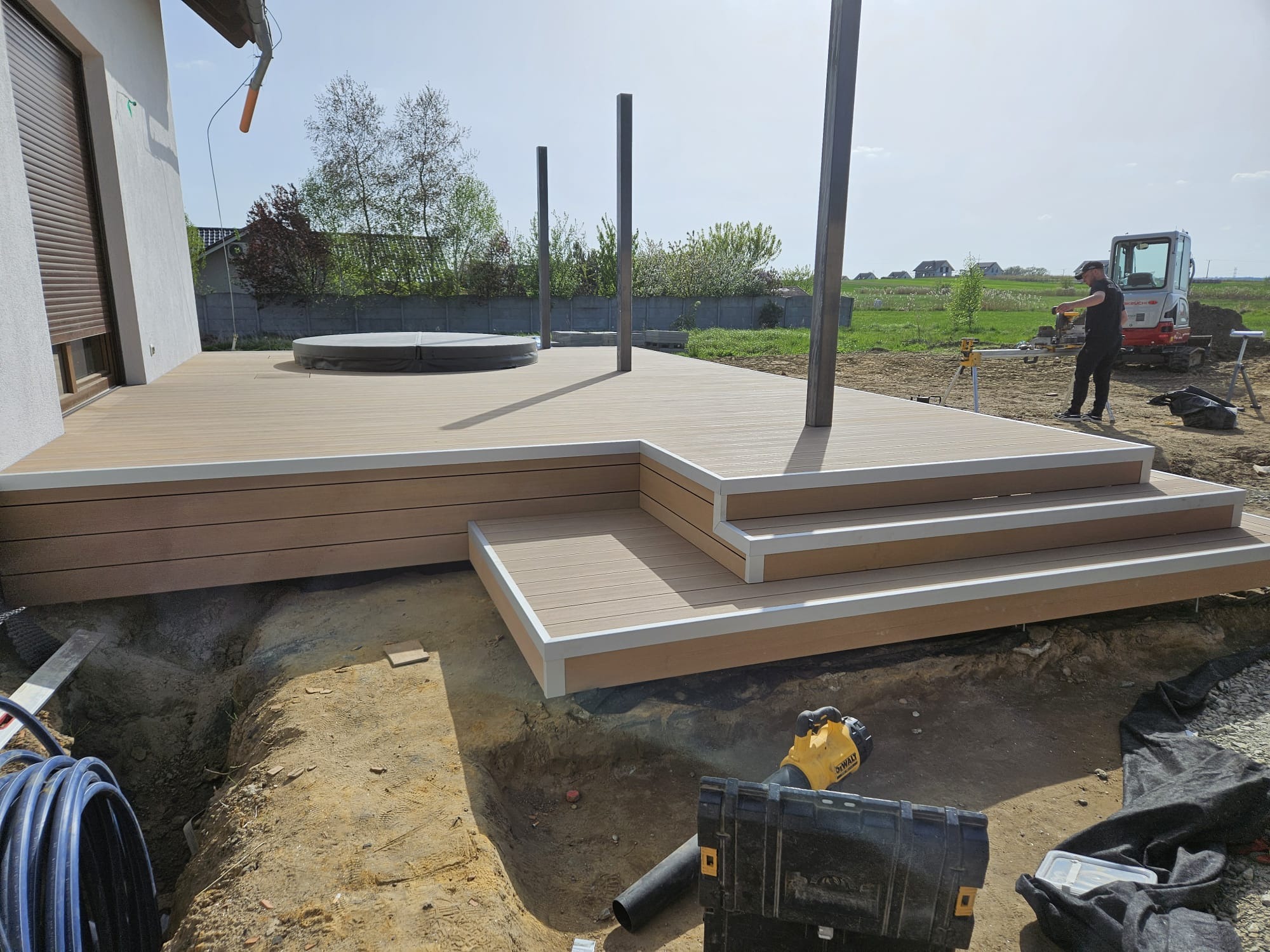 Montaż tarasu z widocznymi słupkami, schodami i przygotowanym podłożem. W tle operator minikoparki.