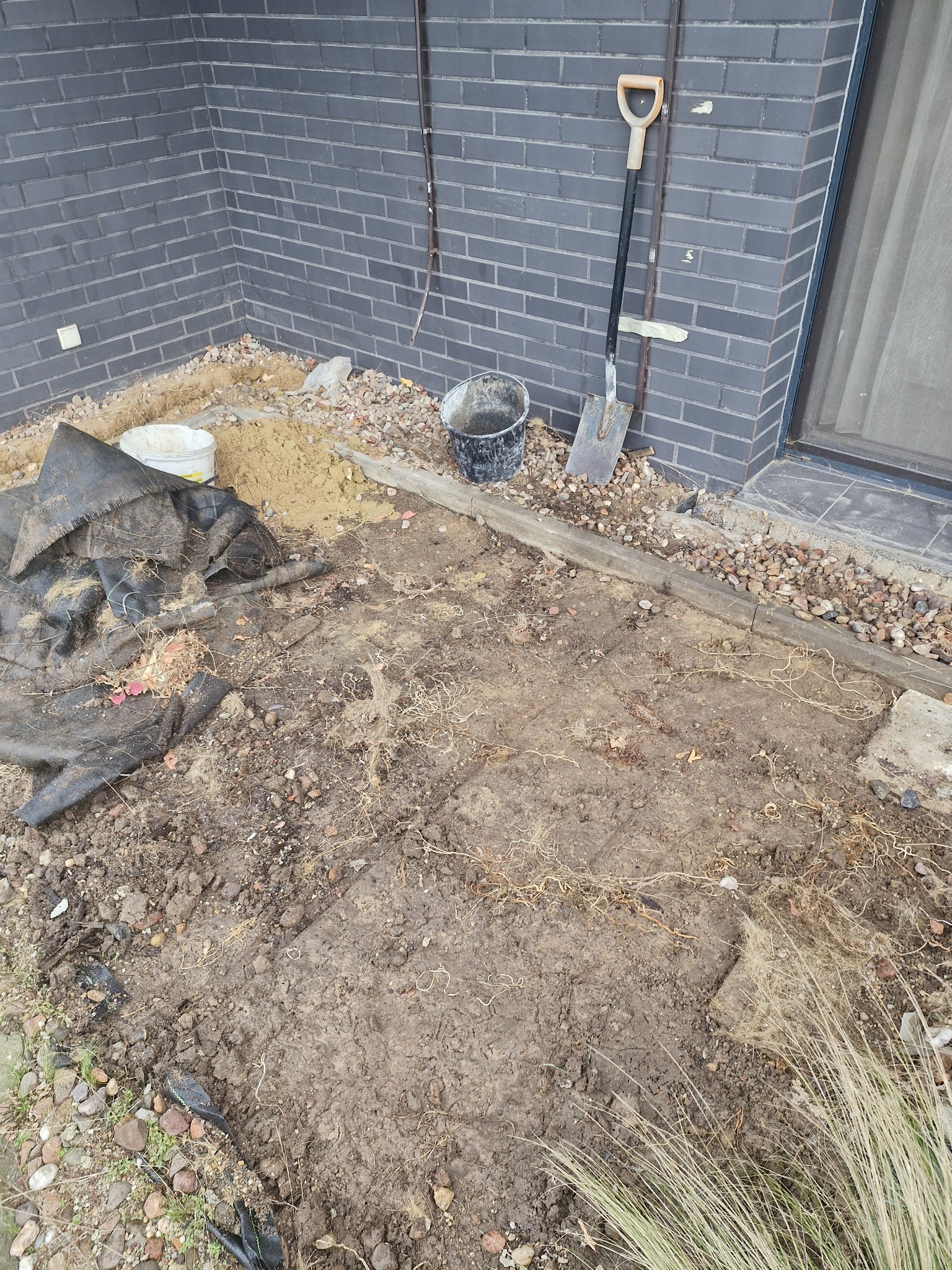 Przygotowanie podłoża pod taras: wyrównany teren z widoczną folią, piaskiem i gruzem, narzędzia oparta o ścianę z cegły.