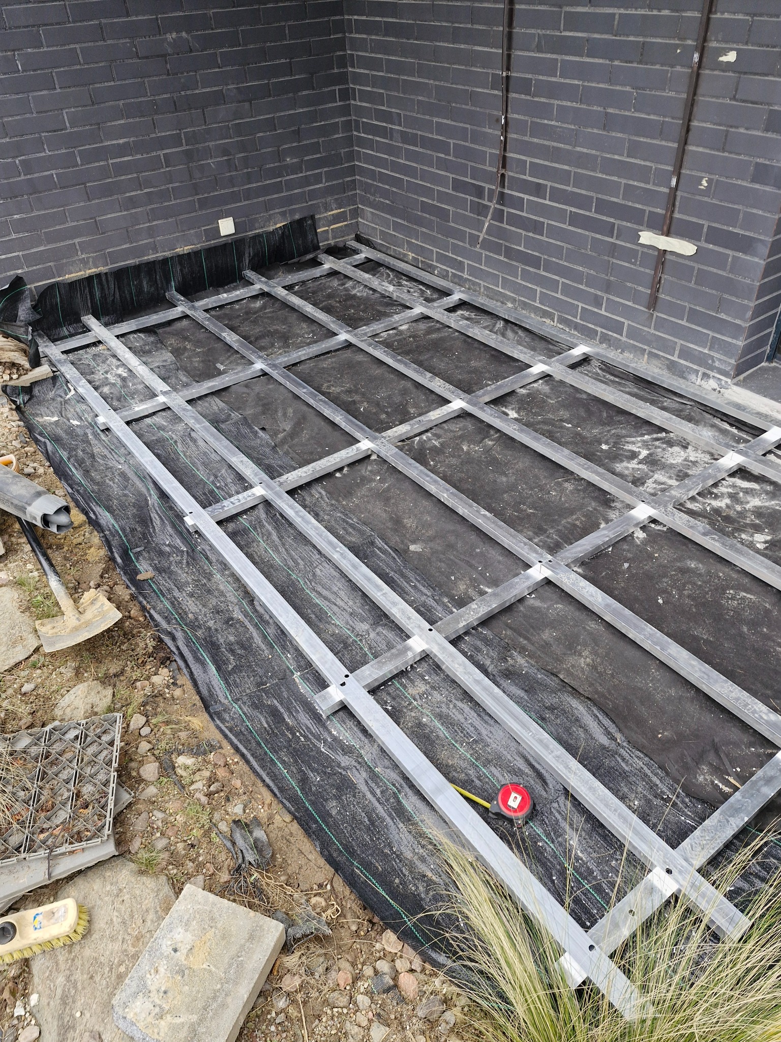 Konstrukcja aluminiowa pod taras na czarnej geowłókninie, widoczna taśma miernicza i narzędzia, ciemna ceglana ściana w tle.