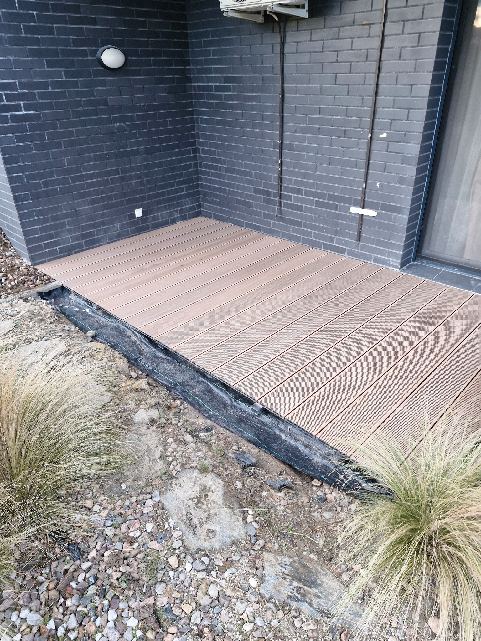 Fragment tarasu z jasnych desek, częściowo ułożonego, z widoczną czarną folią pod spodem, na tle ciemnej, ceglanej ściany budynku i ziemi z kamieniami oraz kępami trawy.