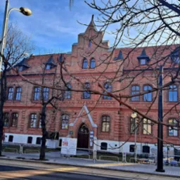 Ceglany budynek sądu lub urzędu z elementami architektury gotyckiej, widziany z ulicy przez gałęzie drzew bez liści.