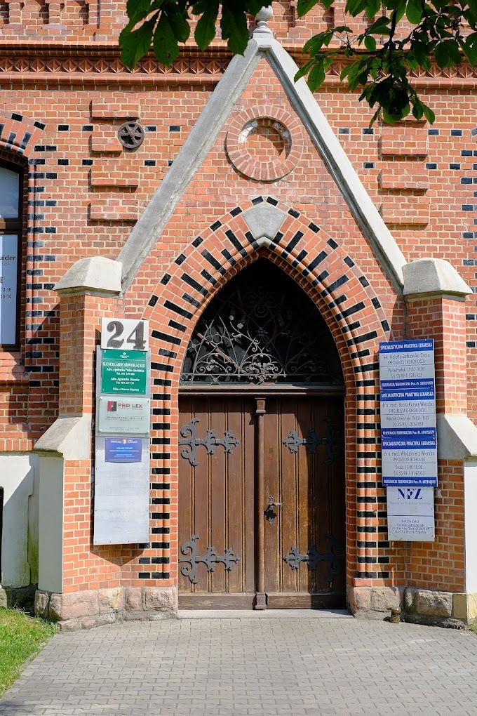 Fasada budynku z czerwonej cegły z gotyckim łukiem nad drewnianymi drzwiami, tabliczki informacyjne po obu stronach wejścia, numer 24 nad wejściem.