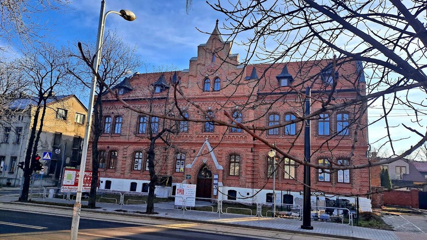 Ceglany budynek sądu lub urzędu z elementami architektury gotyckiej, widziany z ulicy przez gałęzie drzew bez liści.