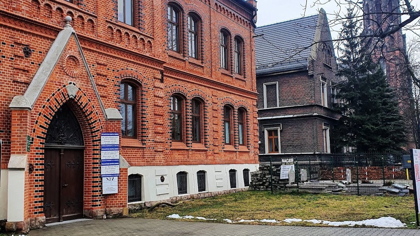 Fasada budynku z czerwonej cegły z tablicami informacyjnymi przy wejściu, obok budynek z ciemnej cegły i fragment kościoła w tle.