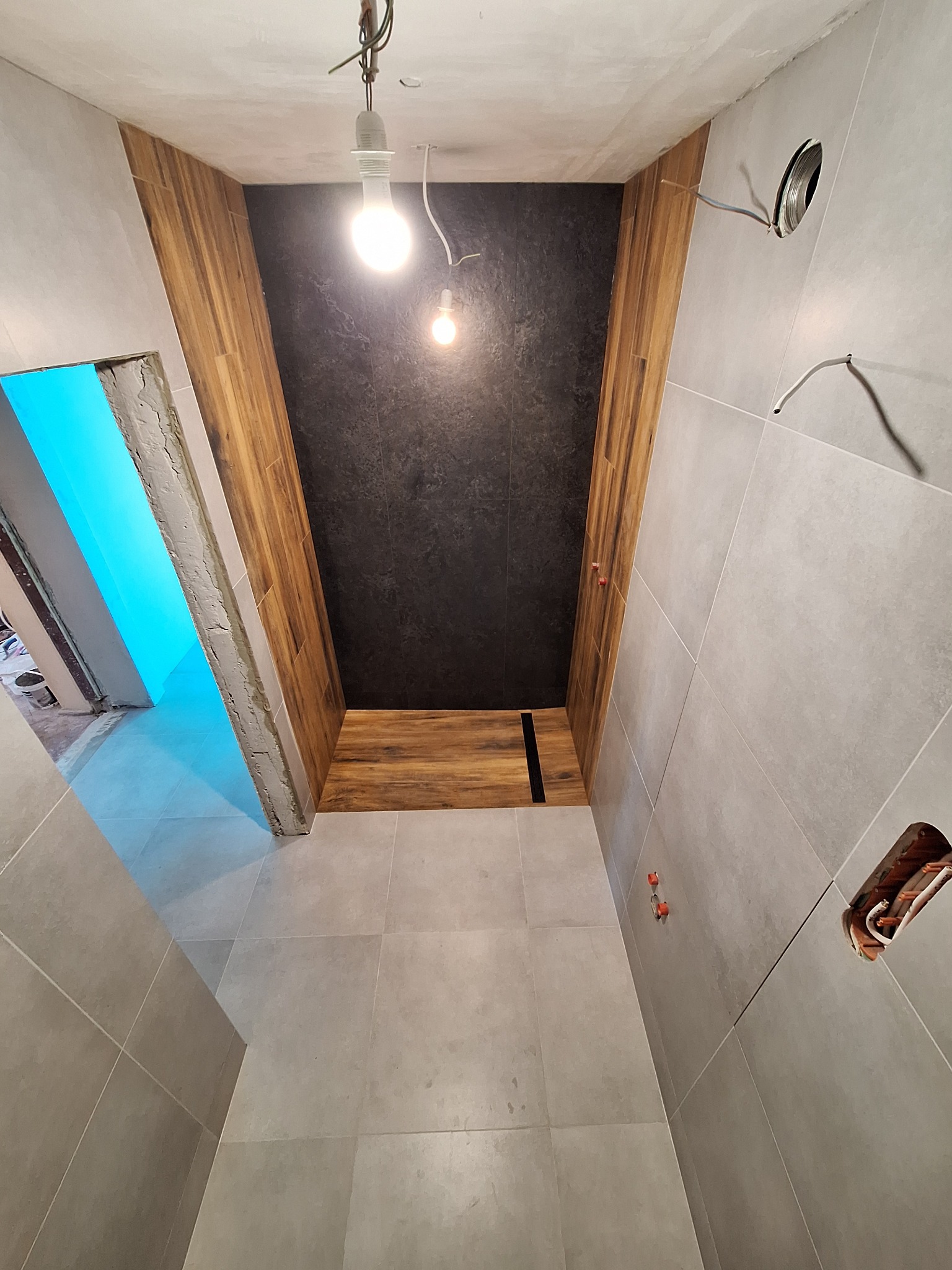 Wnętrze łazienki w trakcie prac wykończeniowych: szare płytki na ścianach i podłodze, drewnopodobna okładzina na ścianie, widoczne instalacje elektryczne i hydrauliczne, surowy stan wykończenia