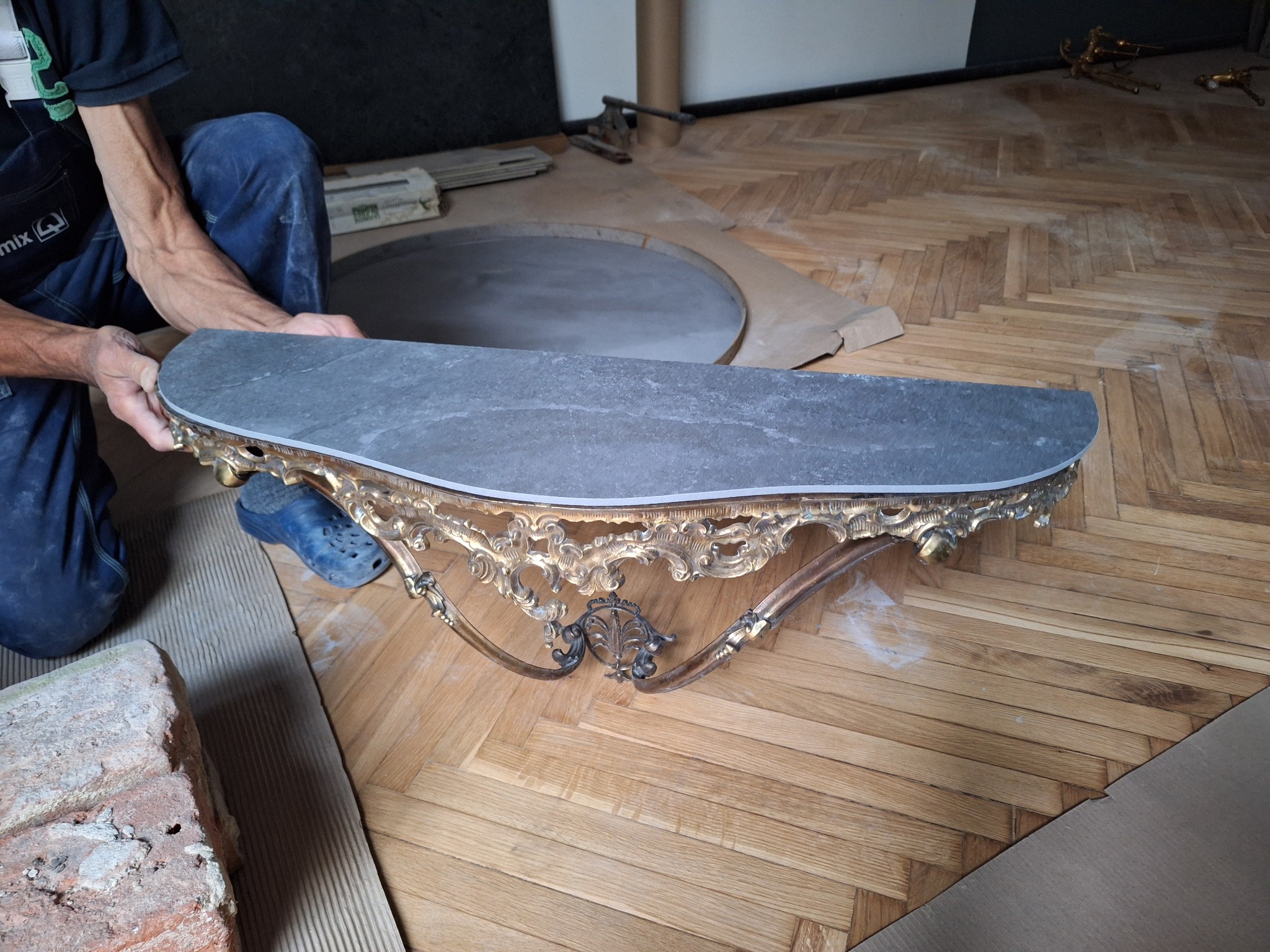 Mężczyzna w niebieskich chodakach trzyma półkę z marmurowym blatem i bogato zdobioną, złoconą ramą na parkiecie w jodełkę, przygotowując ją do montażu w łazience.