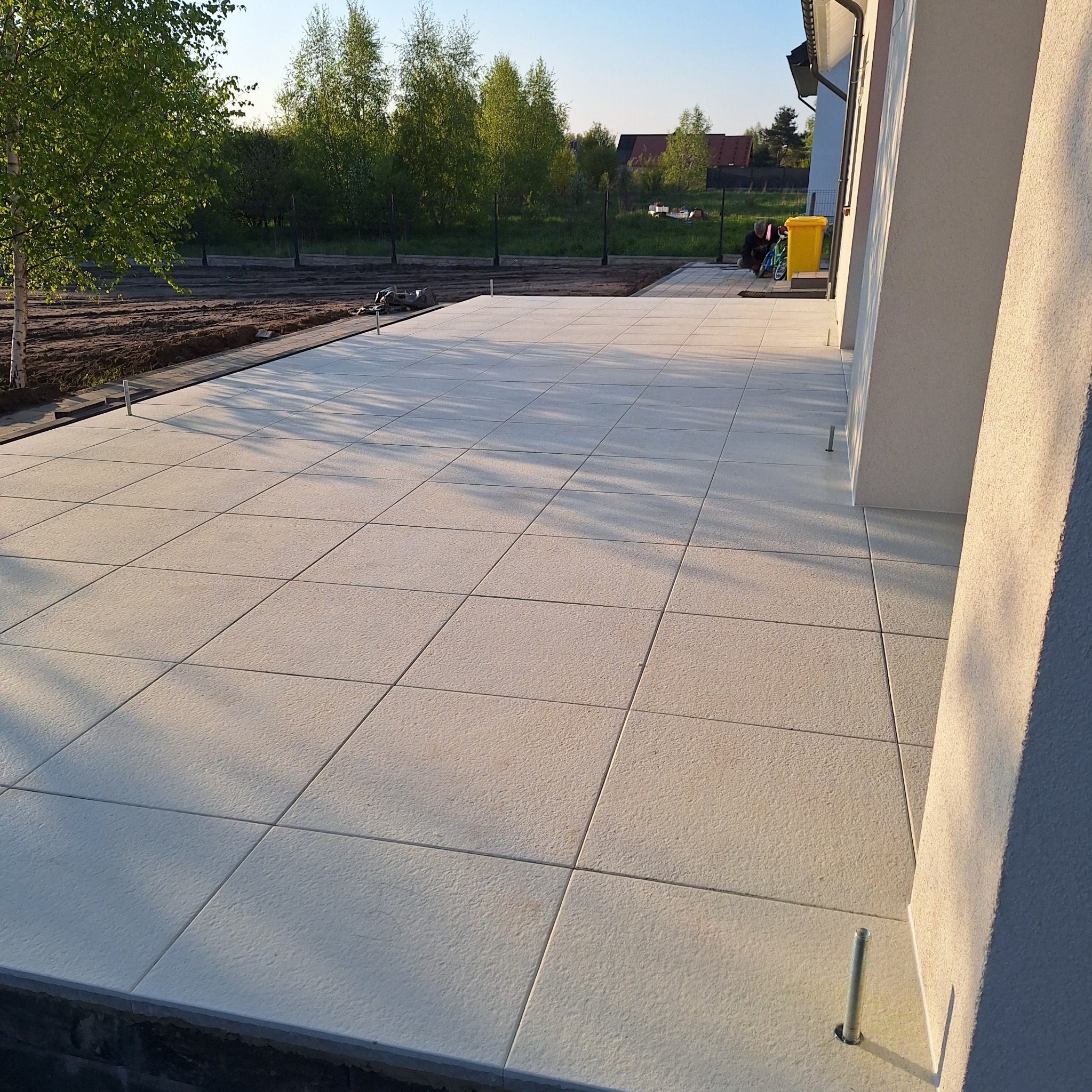 Nowo wyłożony, jasny taras z płyt betonowych, widoczne metalowe trzpienie stabilizujące, w tle trawnik i drzewa.