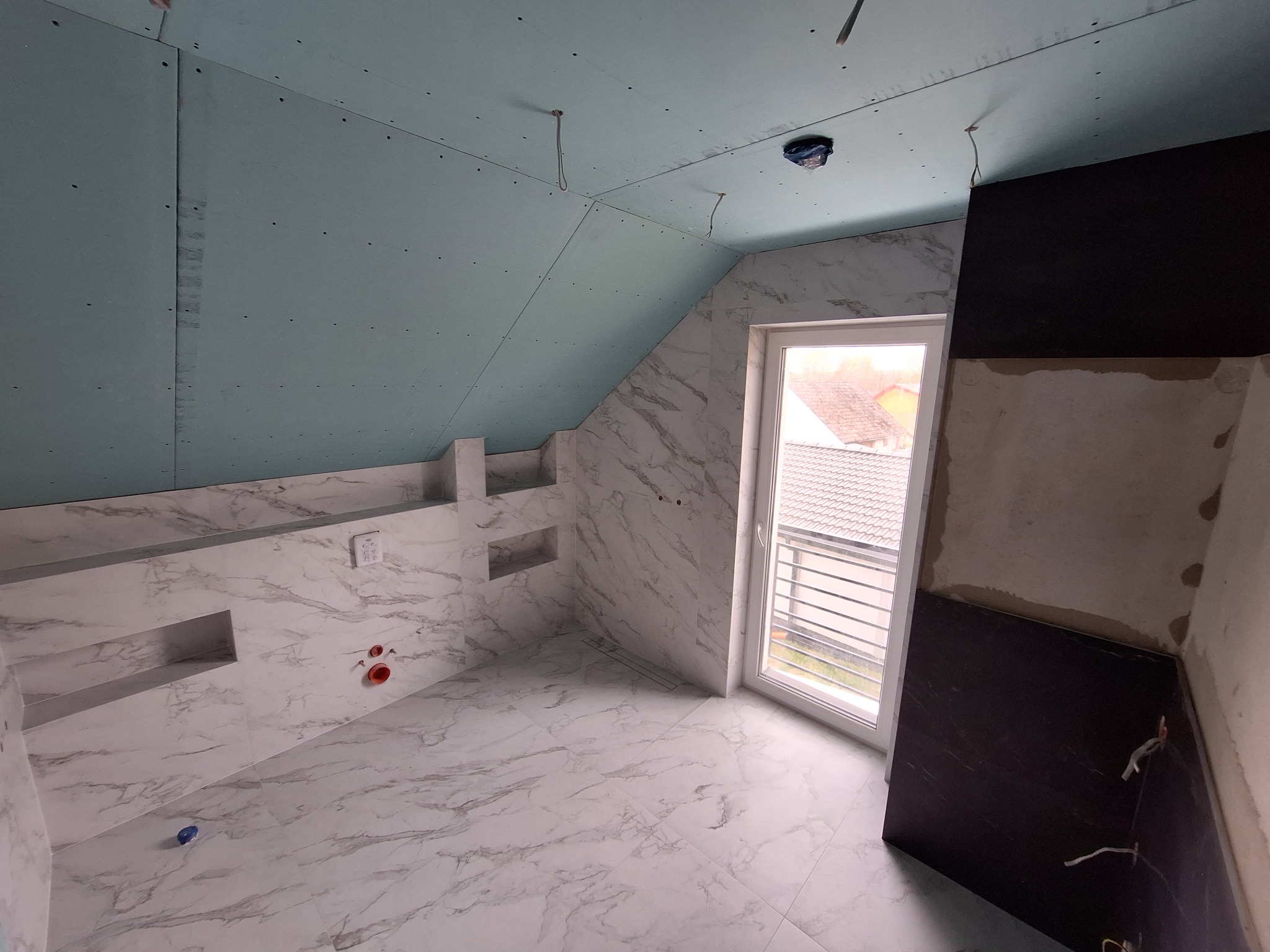 Remontowana łazienka na poddaszu: ściany wyłożone białymi płytkami imitującymi marmur, widoczne wnęki, sufit z płyt gipsowo-kartonowych w kolorze niebieskim, częściowo wykończona ściana z ciemnymi...