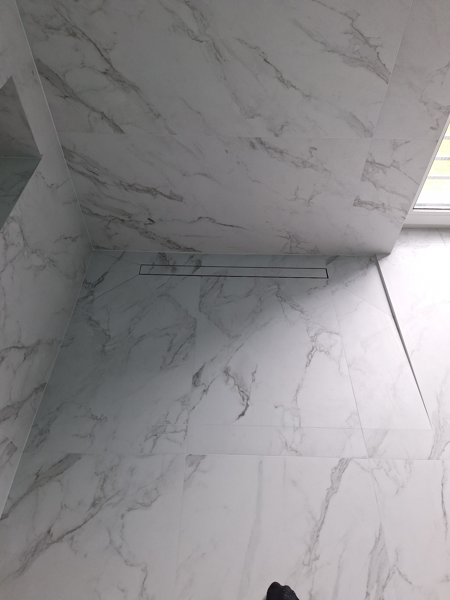 Nowoczesna łazienka z białymi płytkami imitującymi marmur i minimalistycznym odpływem liniowym w podłodze.