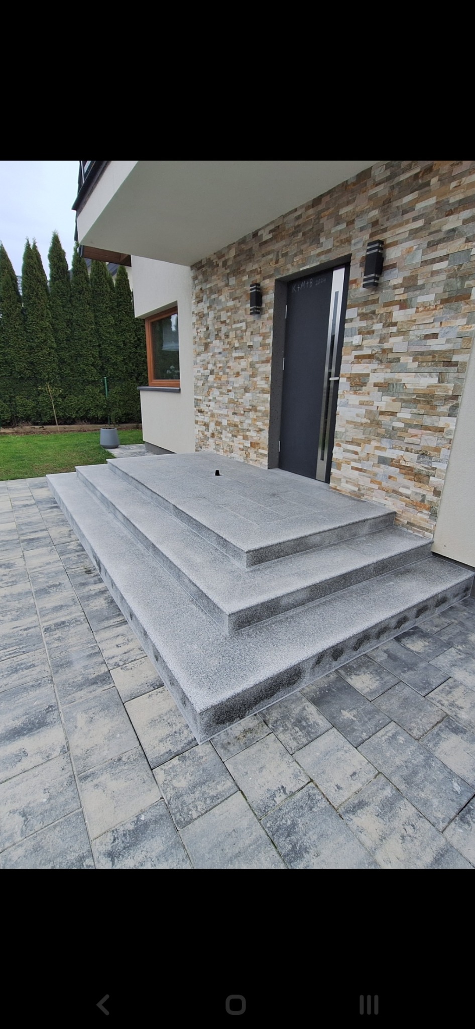 Wykończone granitowe schody wejściowe do domu z elewacją z kamienia dekoracyjnego, czarnymi lampami i drzwiami.