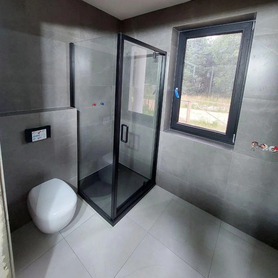 Nowoczesna łazienka z szarymi płytkami, kabiną prysznicową z czarnymi ramami i podwieszaną toaletą. Widoczne okno z widokiem na zewnątrz.