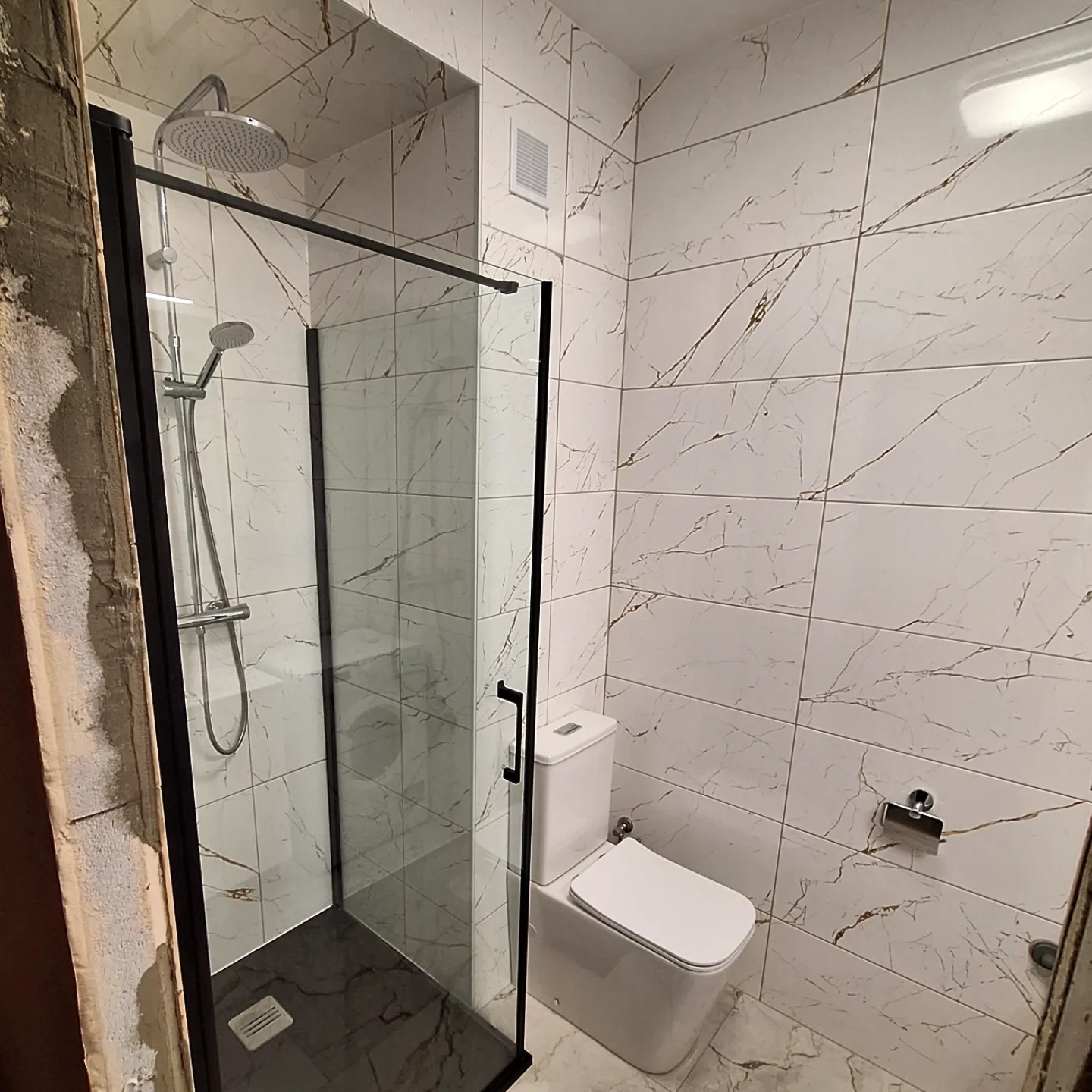 Nowoczesna łazienka z kabiną prysznicową typu walk-in z czarnymi ramami, białą toaletą i płytkami imitującymi marmur Carrara z delikatnymi złotymi żyłkami na ścianach i podłodze, widoczny fragment...