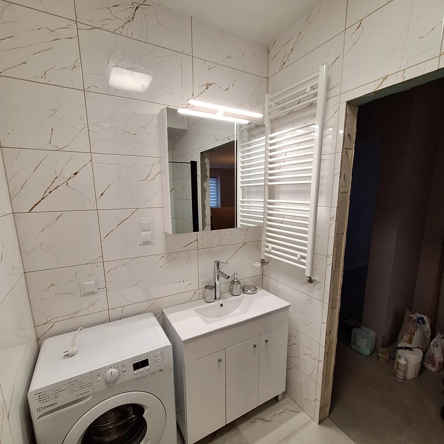Biała łazienka z marmurkowymi płytkami, pralką Indesit, szafką z umywalką i grzejnikiem drabinkowym, widok na niedokończone pomieszczenie obok.