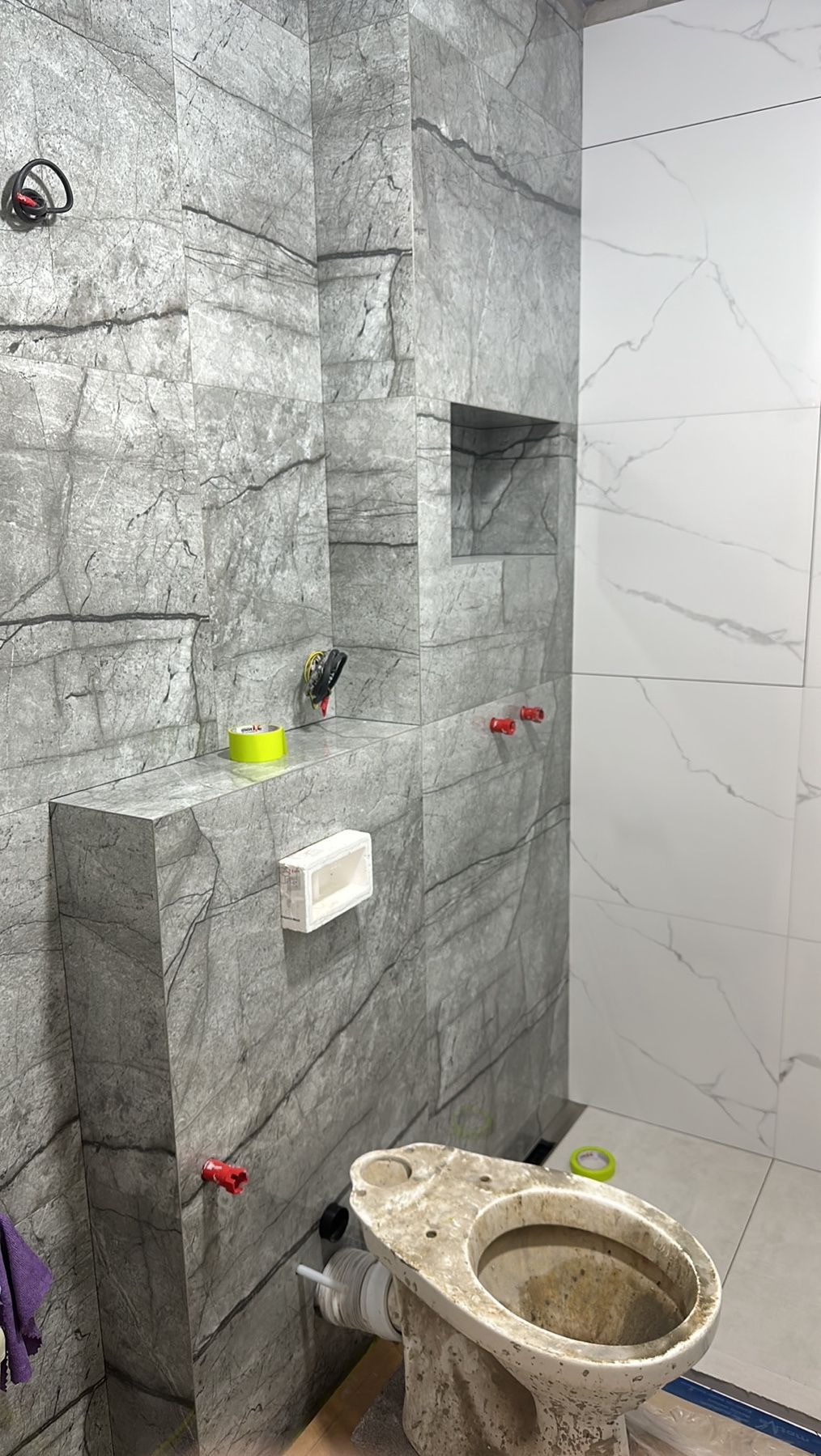 Wnętrze łazienki w trakcie remontu: ściany wyłożone płytkami imitującymi marmur, częściowo ułożone białe płytki, surowy sedes, widoczne narzędzia i materiały budowlane, instalacja hydrauliczna...