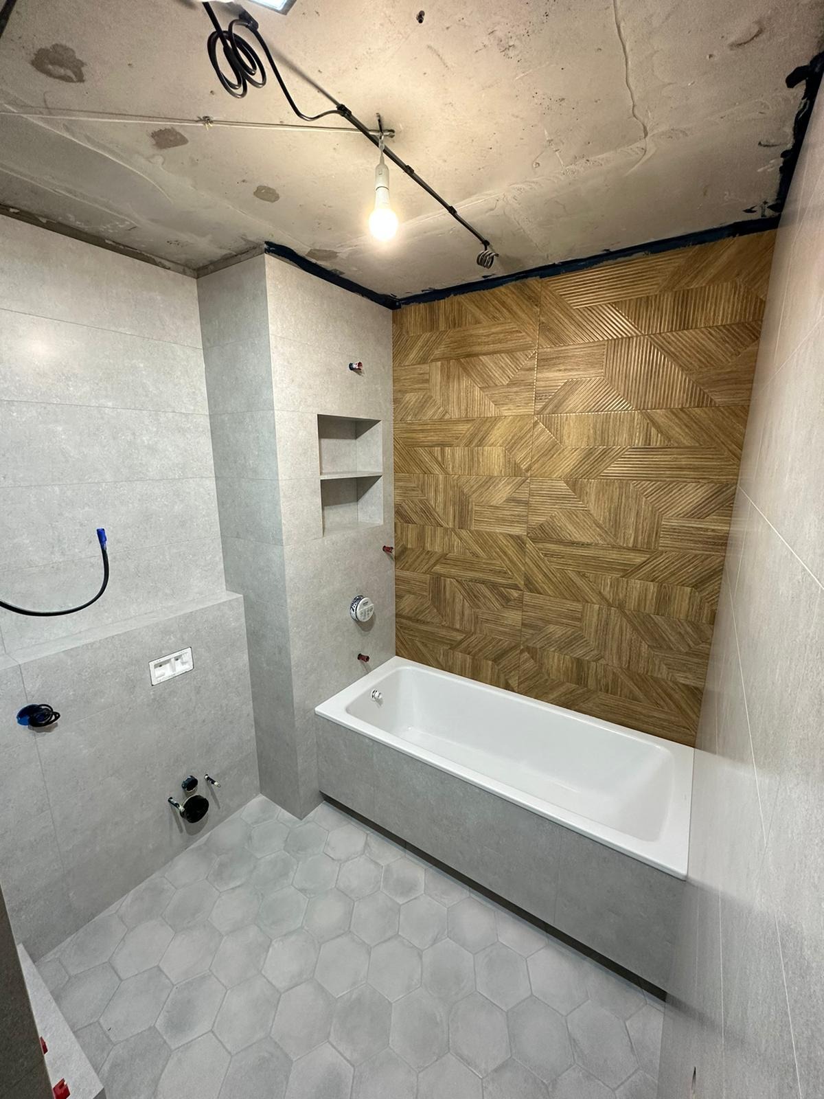Łazienka w trakcie remontu z szarą armaturą, sześciokątną podłogą, drewnopodobną ścianą dekoracyjną, białą wanną, surowym betonowym sufitem i widoczną instalacją elektryczną.