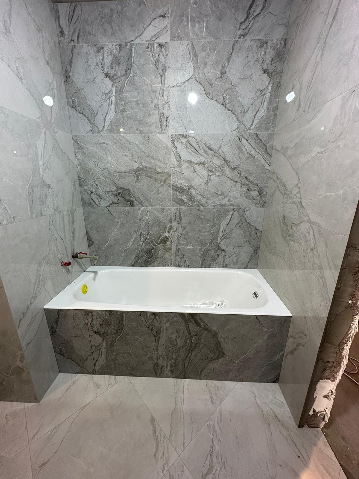 Wykończona łazienka z wanną obudowaną płytkami imitującymi marmur, ściany i podłoga również wyłożone płytkami w podobnym stylu, widoczny fragment niezakończonej ściany po prawej stronie.