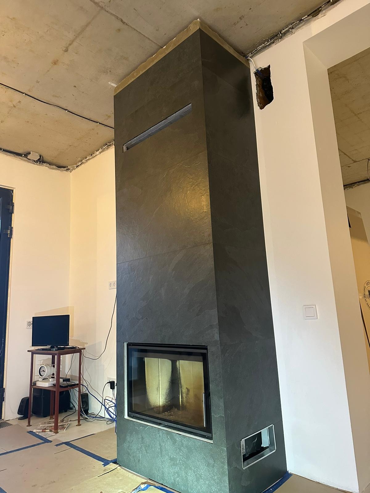 Nowoczesny kominek obłożony płytkami imitującymi łupek, z prostokątnym otworem wentylacyjnym u góry, w surowym wnętrzu w trakcie wykończenia.