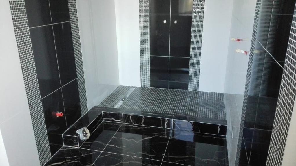 Nowoczesna łazienka z czarnymi marmurowymi płytkami na podłodze i ścianach, mozaika w odcieniach szarości, widoczne elementy instalacji wodnej.