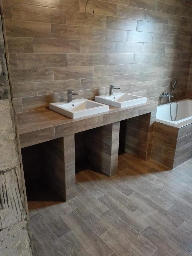 Ukończona łazienka z dwoma umywalkami nablatowymi, wykończona płytkami imitującymi drewno, z widoczną wanną i fragmentem surowej ściany z lewej strony.