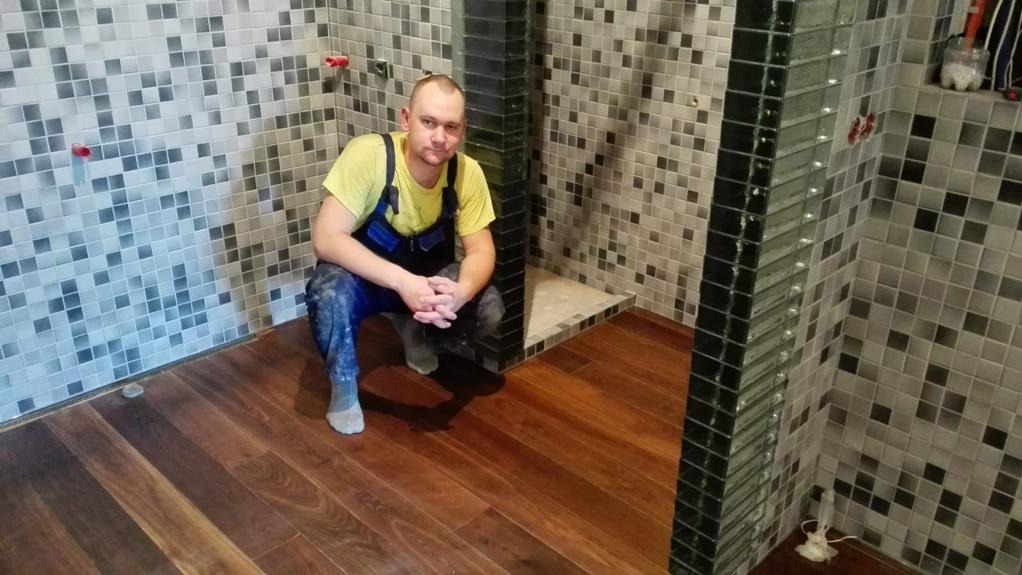 Mężczyzna w roboczym ubraniu kuca w nowo wykończonej łazience z mozaikowymi płytkami na ścianach i drewnianą podłogą, widoczny fragment kabiny prysznicowej z czarnymi płytkami.